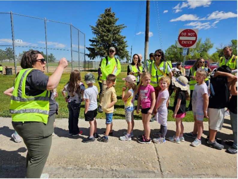 Elementary school students line up on a sidewalk with adults wearing yellow safety vests